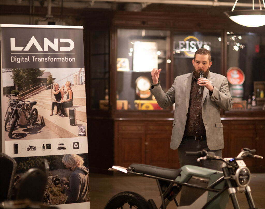 BRITE CEO speaking at a BRITE event standing in front of a LAND moto e-bike